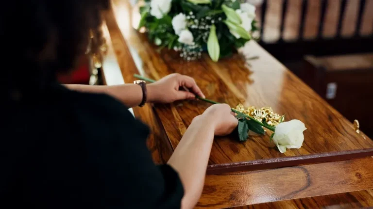 Person in black placing a single white rose on a polished wooden coffin with floral arrangement.