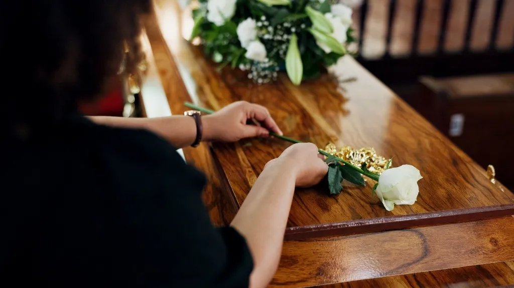 Person in black placing a single white rose on a polished wooden coffin with floral arrangement.