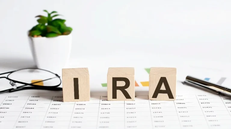 Wooden blocks spelling "IRA" on a financial spreadsheet with eyeglasses, pen, and potted plant.