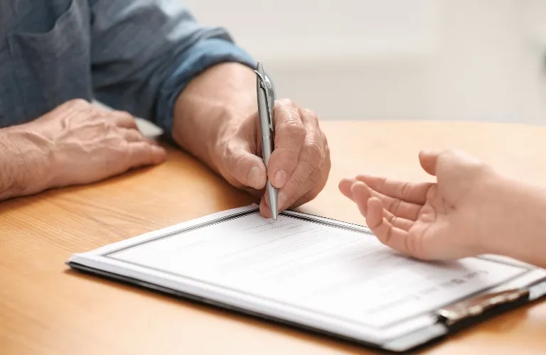 Two individuals at a wooden table, one holding a pen poised to sign a document on a clipboard.