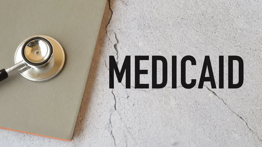 Stethoscope on closed gray folder beside the word "MEDICAID" on cracked concrete surface.