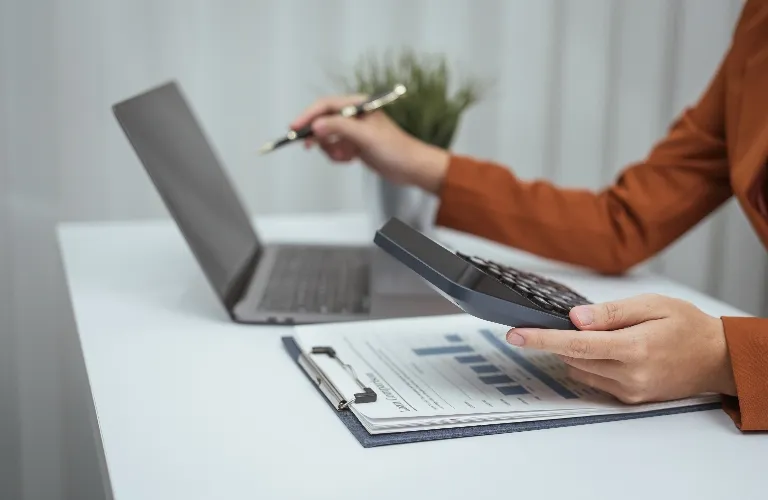 Person using calculator and pointing at laptop screen with pen, documents on desk.