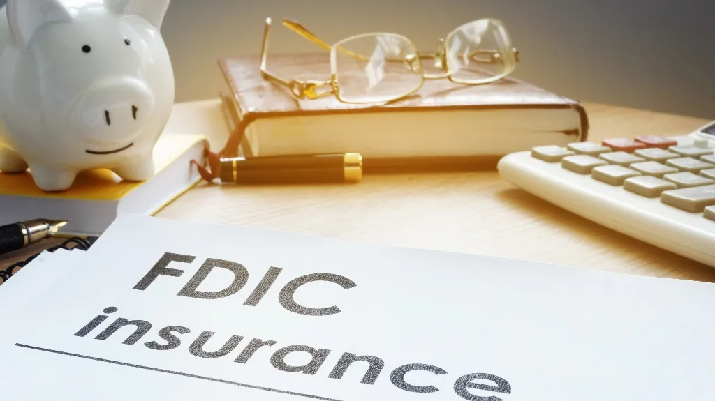 Document titled "FDIC insurance" on desk with piggy bank, glasses, pen, and calculator.