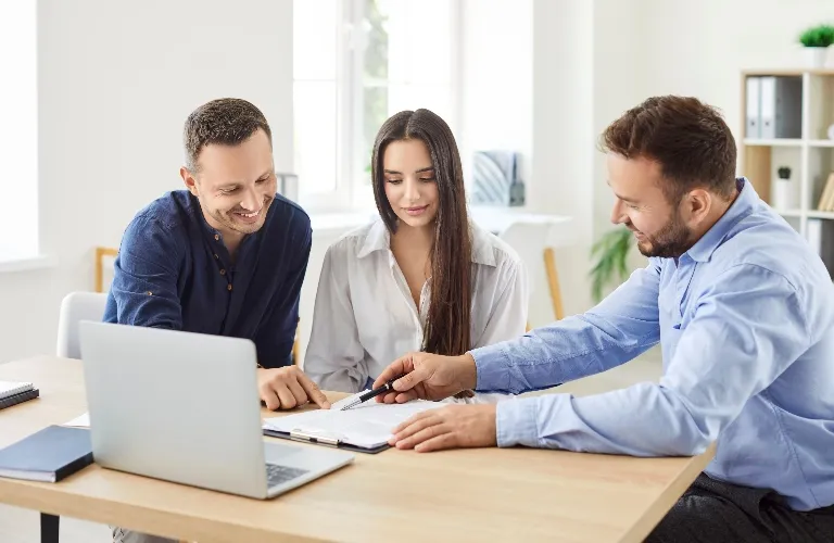 Three professionals reviewing documents together at a light wooden office desk.