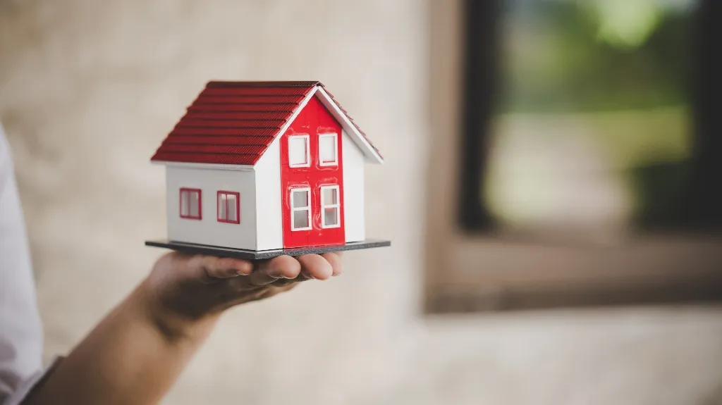 Hand holding a small white and red model house with a tiled roof.