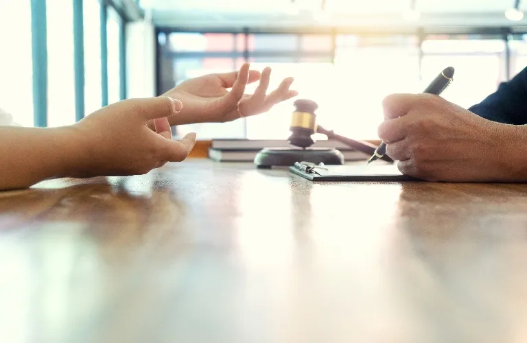 Hands of two individuals at a wooden table, one writing on a clipboard, with a judge's gavel in the background.