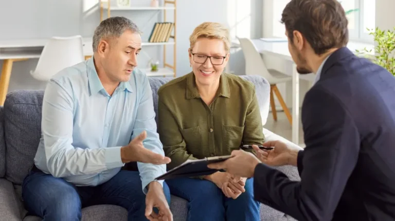 Three professionals engaged in a discussion, with a man showing a clipboard to a seated couple.