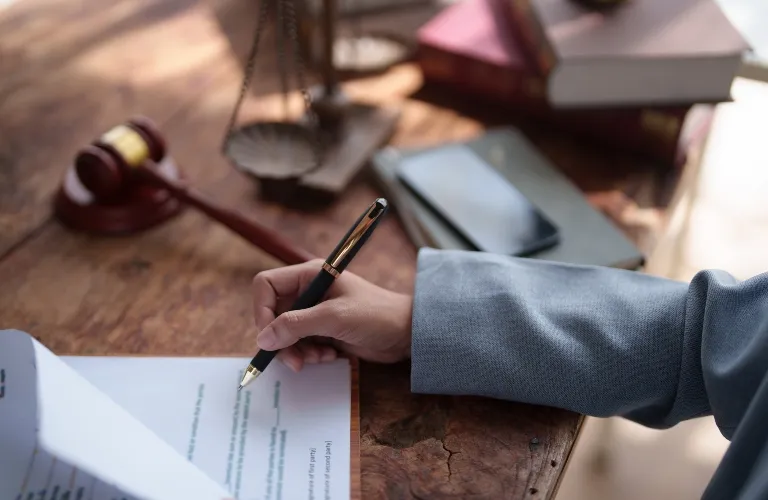 Person in gray suit writing on document with pen on wooden desk, gavel and scales in background.