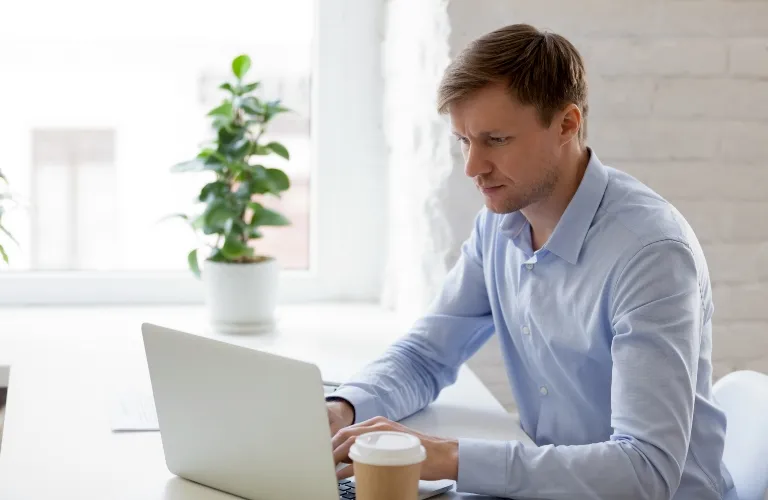 Professional man in light blue shirt working on laptop at bright desk with coffee cup and plant.