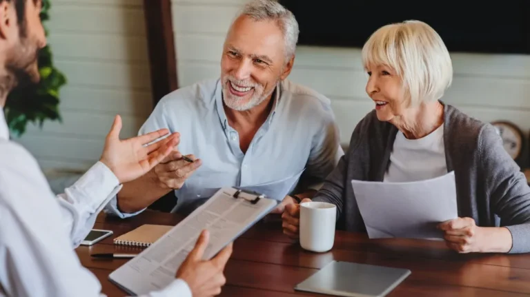 Three adults, two elderly and one younger, engaged in discussion with documents and a tablet at a wooden table.