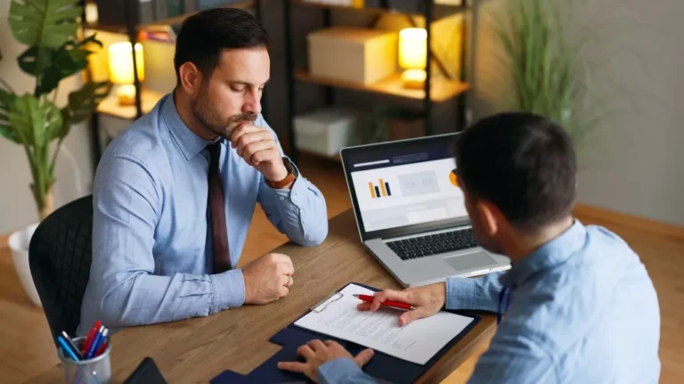 Two men in blue shirts engaged in a business discussion with laptop and documents.